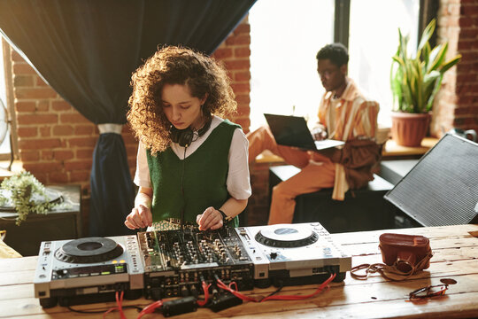 Young woman in casualwear standing by wooden table and using dj controller while creating new music at leisure against black man