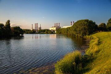 The landscape of a city park with a river and green trees, industrial production pipes in the background.