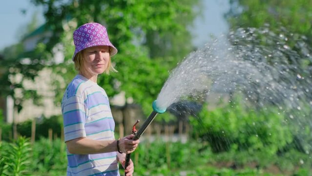 Happy female farmer watering her garden with hose in sunny summer day. Positive adult woman in funny hat pouring green plants in backyard.