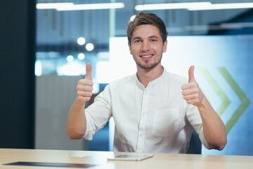 Man in modern office working, talking on video call looking at camera smiling showing thumbs up