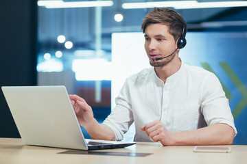 A young office worker working with a laptop uses a headset for video communication, a man from the support service communicates online with the client