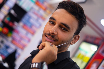 Handsome barber arab man smiling for camera and showing hairdressing scissors while working in vintage salon against barbershop background - obrazy, fototapety, plakaty