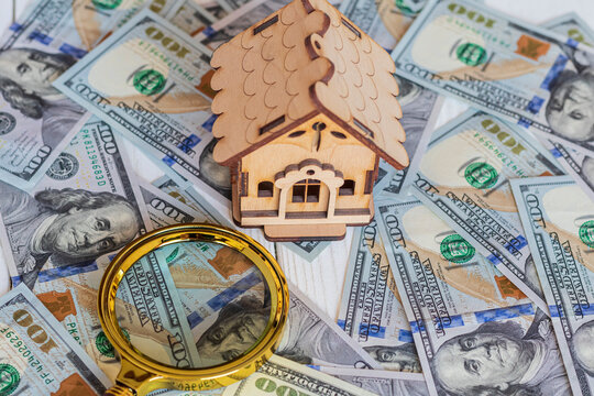 A toy house made of wood with a roof costs money, on a hundred-dollar bill, next to a magnifying glass. Concept: mortgage, purchase, sale of real estate, rental housing - Powered by Adobe