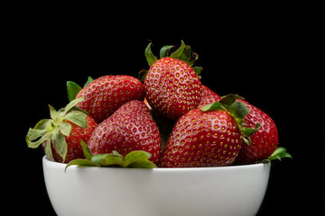 Ripe red strawberries with green tails in a white plate on a black background. Sweet dessert of fresh berries on the table.