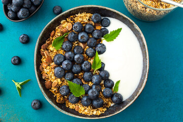 Homemade granola with yogurt, blueberries and mint leaves in a bowl on a turquoise background, delicious healthy breakfast