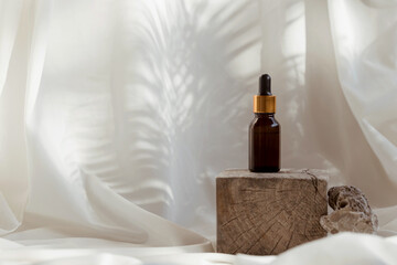 Amber glass bottle of essential oil or serum on white fabric background with wooden podium shadows. Natural skincare cosmetic concept