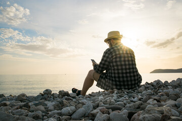 Rear side view of man with smartphone sitting on beach terrace on sunny day outside background with copy space - vacation or freelance or social network concept