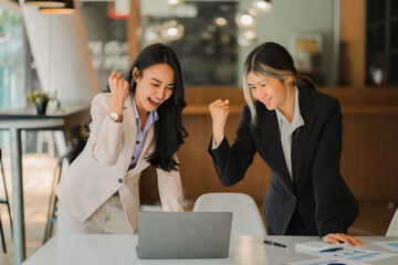 Two Asian business women rejoice at their success in business and raise their hands in front of their laptop computers. Colleagues rejoiced at the victory of the group. - obrazy, fototapety, plakaty