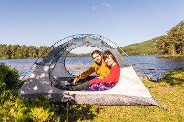 Photo of a couple sitting inside a tent after spending the night camping in front of a lake in...
