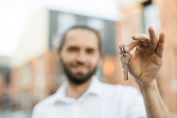 Focus on hand with keys. Handsome young businessman in white shirt holding keys of new flat, looking at camera and smiling, standing near the building in the middle of the street.