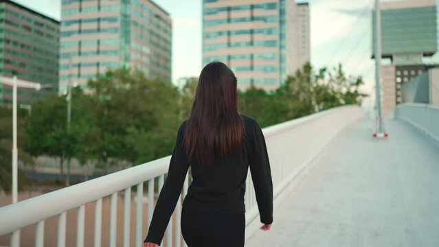 Young woman loose hair wearing black sports hoodie is walking fast across bridge modern city background. Back view