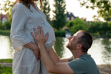 Naklejka na ściany i meble Beautiful pregnant young woman outside, relaxing and enjoying life in nature, warm sunny picture. Lovely couple near the pond. Maternity photoshoot. Young man standing with his pregnant woman