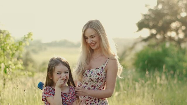 Smiling blonde mom in a summer dress combs her daughter's hair. Daughter and mom laugh and have fun together outdoors