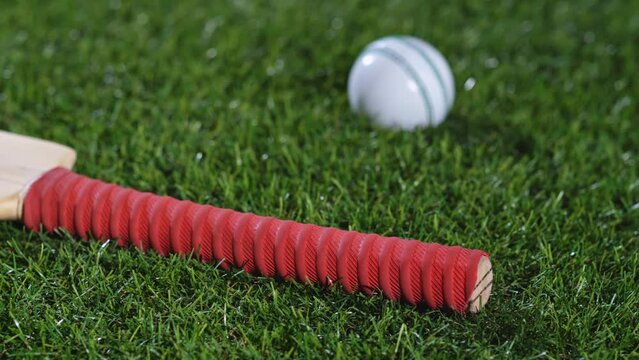 A low angle slider shot along a wooden cricket bat with a red grip with white and green cricket balls sitting in the grass.