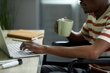 Side view closeup of African American man in wheelchair using laptop while working from home and drinking coffee