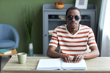Front view portrait of adult black man reading tactile book in braille and wearing dark sunglasses,...