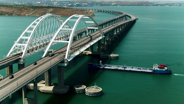 A ship passes under a large bridge with moving cars. Shot. Aerial view of a bridge and a ship with turquoise water, time lapse effect.