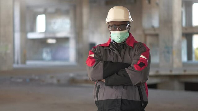 Slow Motion African american worker stands at construction site, looks at the camera and takes off medical protective mask, but unsuccessfully and funny, mask caught on glasses, worker smiling