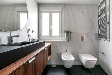 Modern design bathroom with white porcelain toilets, wooden furniture, black porcelain sink, circular mirror with bluetooth, chrome towel radiator and black marble floors