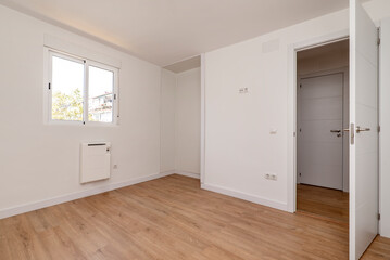 Empty room in a renovated apartment with white painted walls, white woodwork and oak colored parquet floor, with space for a wardrobe and aluminum window with sliding leaves