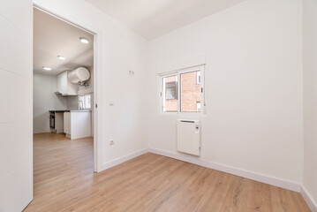 Empty room in renovated apartment with white painted walls, white joinery and oak parquet floor and aluminum window with sliding panels and electric radiator below and access to a kitchen