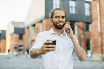 Portrait of handsome caucasian man having paper cup of fresh beverage talking to partner using smartphone walking on city street looking at camera.