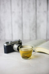 Brewed herbal green tea in a transparent cup next to an open book and a vintage camera on a light background