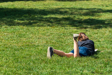 A school-age girl reads a book while lying on the grass in a city park on a sunny Sunday afternoon. Children's self-education in their free time, the importance of reading