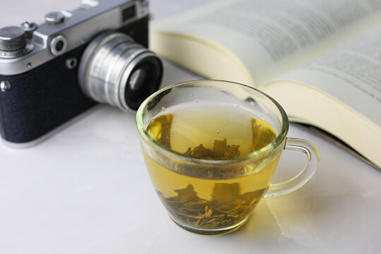 Brewed herbal green tea in a transparent cup next to an open book and a vintage camera on a light background