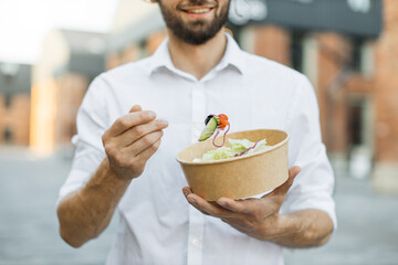 Close up view of hand of businessman having a vegetables salad for lunch, healthy eating standing on background of city, lifestyle concept, unrecognizable person.