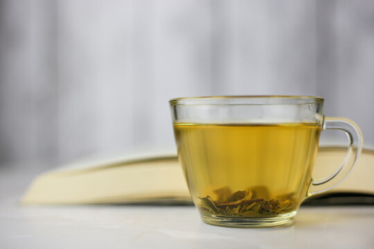 Brewed herbal green tea in a transparent cup in front of an open book on a light background.