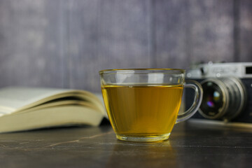 Brewed herbal green tea in transparent cup near open book and vintage camera on dark background