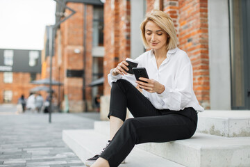 Business woman in white shirt talking on smartphone. Professional businesswoman using mobile phone smiling drinking coffee, sitting on stairs near office building in city.