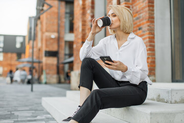 Cheerful young blond woman wearing white shirt and black pants sitting on stairs outdoors, drinking takeaway coffee cup, using mobile phone.