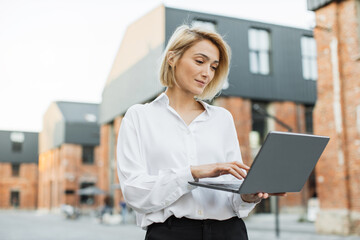 Stylish business female using modern laptop for remote work while walking on the street near office building. Smiling woman freelancer enjoying favorite work during break.