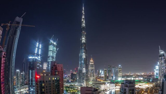 Panorama of Dubai Downtown cityscape with tallest skyscrapers around aerial during all night timelapse. Construction site of new towers and busy roads with lights turning off from above