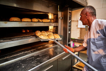 Professional baker in uniform takes out a cart with freshly baked bread from an industrial oven in...