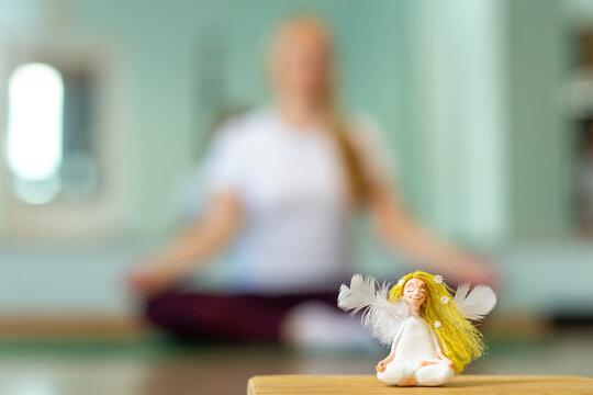 A doll in the form of an angel on blur background with healthy female practicing yoga indoor.