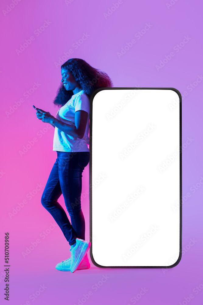Poster full length of young black lady using mobile device, standing near huge cellphone with mockup for de