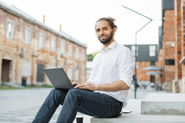 Successful business man dressed in white shirt and jeans sitting on stairs outdoors and typing on modern laptop. Attractive freelancer using portable computer for remote work.