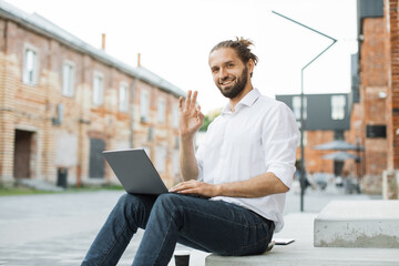 Successful business man dressed in white shirt and jeans sitting on stairs outdoors and typing on modern laptop. Attractive freelancer using portable computer for remote work showing sign okay.