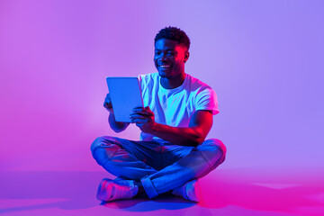 Handsome black guy using tablet computer, sitting cross legged in neon light