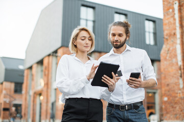 Attractive satisfied business couple standing outdoors near office building and making online payment using credit card, tablet and smartphone. Checking account balance, buying of goods concept.