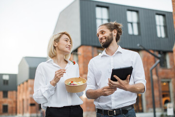 Charming woman holding a cardboard bowl with fresh organic salad and attractive man with tablet...