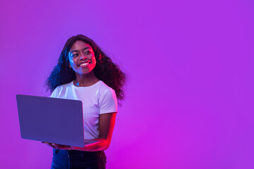 African American lady using laptop for online work, education or communication, looking at empty space in neon light - obrazy, fototapety, plakaty