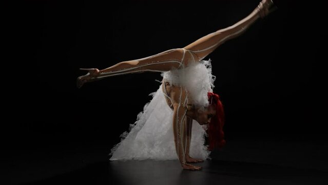 Side view of flexible slim woman in stage costume doing back walkover standing up looking at camera. Wide shot portrait of confident seductive Caucasian gymnast posing performing at black background