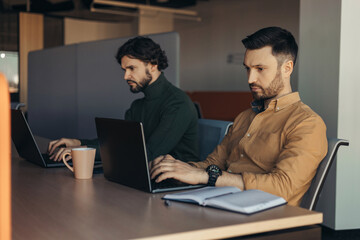 Fototapeta na wymiar Focused male colleagues using laptop computers at desk in modern office, free space