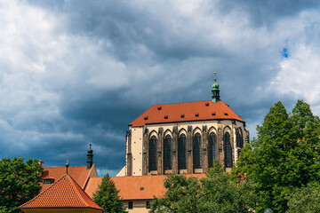 Obraz premium Golden crosses on the orange roof of an old gothic temple under a cloudy rainy sky behind the green crowns of large trees in Prague, Czech Republic. Tourist travels in European cities