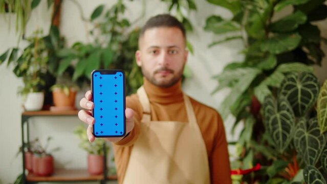 Forist holding phone with chroma key. Greenhouse worker portrait. Arabian man botanist smiling, small business owner. Gardener working in flower shop, plant store. 