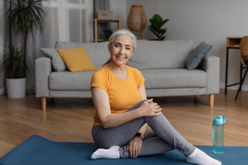 Healthy lifestyle concept. Happy senior woman sitting on sports yoga mat at home in living room and...